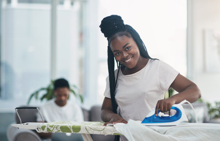 Hubby handles business, I handle the housework. a young woman ironing clothing with her husband on the sofa in the background.の写真素材