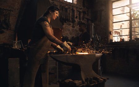 The steel world has much to thank for her work. a young woman hitting a hot metal rod with a hammer in a foundry.の写真素材