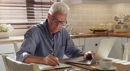Scheduling meetings even at home. a senior man sitting alone in the kitchen and using his laptop to work from home.の写真素材