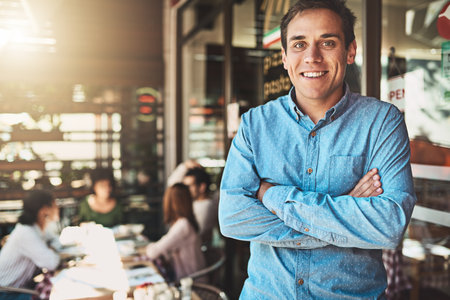 He calls the shots at work. Portrait of a confident young man standing with his arms folded inside of a coffeeshop during the day.の写真素材