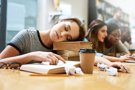 Prepping for finals got her like...a young woman looking tired while studying in a college library.