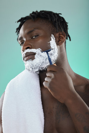 Black man, studio and cream for shaving, beauty or razor for facial, cosmetics or skincare by blue background. African gen z model, hair removal foam and shave for aesthetic, cleaning and self careの写真素材
