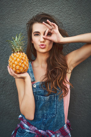 I only see pineapples today. Portrait of a carefree young woman showing a hand gesture on her face while holding a pineapple against a grey background.の写真素材