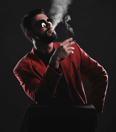 Fashion, smoke and red with a man model in studio on a dark background wearing a suit for style. Smoking, mafia and edgy with a handsome young male posing on black space holding a cigaretteの写真素材