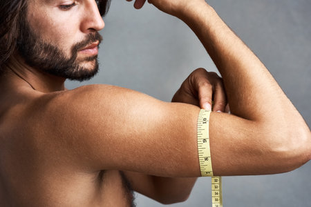 Building the biceps of his dreams. Studio shot of a handsome young man measuring his bicep against a grey background.の写真素材