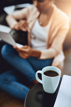 Home is where its at. a cup of coffee on a table with a woman reading a document in the background at home.の写真素材