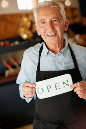 Welcome, weve been waiting for you. Portrait of a senior man holding up a sign on opening day of his small business.の写真素材