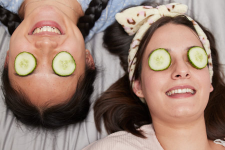 Friends, facial and skincare with cucumber by women relax, spa day and bonding while lying on a bed...