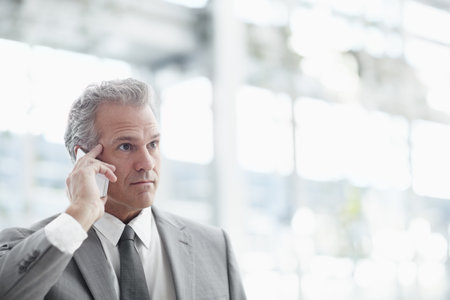 Keeping in touch with junior partners. Mature businessman using a cellphone while standing in the office.の写真素材