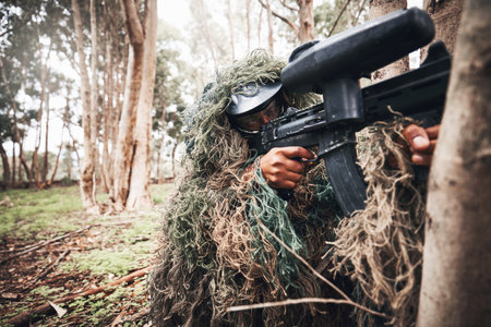 Paintball gun, forrest and man with aim by trees for outdoor war game, strategy or focus in natural camouflage. Shooter, sniper and helmet for safety with eyes on target for winning in military gamesの写真素材