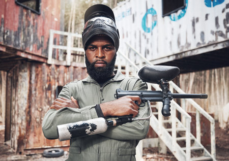 Arms crossed, portrait and black man with a paintball gun for fitness, training or a game. Exercise, focus and serious African player with a rifle for action, military work and outdoor competitionの写真素材