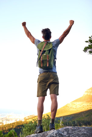 He made it. a handsome young man standing with his arms raised while hiking.