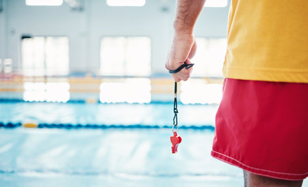 Hands, lifeguard and whistle by swimming pool for water safety, security or ready for rescue indoors. Hand of expert swimmer holding signal tool for warning, safe swim or responsibility for awarenessの写真素材