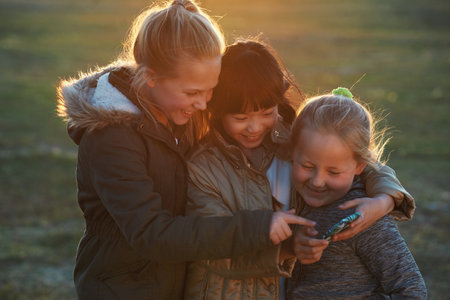 Friends, girls outdoor and smartphone for connection, online games and happiness on summer break. Young people, female children and kids with cellphone, bonding and watching funny videos on fieldの写真素材