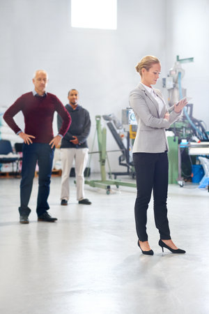 Assuring quality on the shop floor. A manager using a tablet during a factory inspection with her coworkers in the background.の写真素材