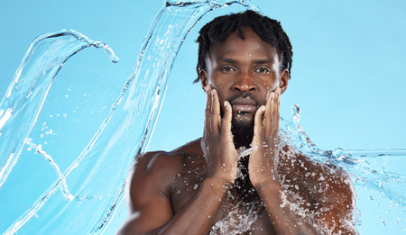Water splash, facial skincare and portrait of black man with hands on face, morning cleaning isolated on blue background. Hygiene, male model and grooming for health, wellness and skin care in studioの写真素材