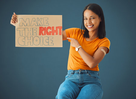 Black woman, portrait and pointing at protest poster, stop covid or vaccine opinion in healthcare. Happy gen z girl, billboard or wellness choice with plaster, cardboard sign and safety by blue wallの写真素材