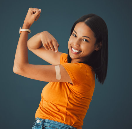 Vaccine, plaster and portrait of a woman in a studio with a strength gesture after being vaccinated. Happy, smile and proud female model with a vaccination band aid isolated by a dark blue backgroundの写真素材