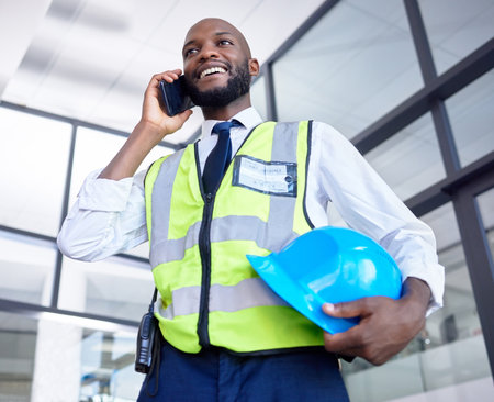 Black man, construction worker or phone call in building planning, networking or industrial property design in low angle. Smile, happy or engineering architect with mobile technology, helmet or ideasの写真素材