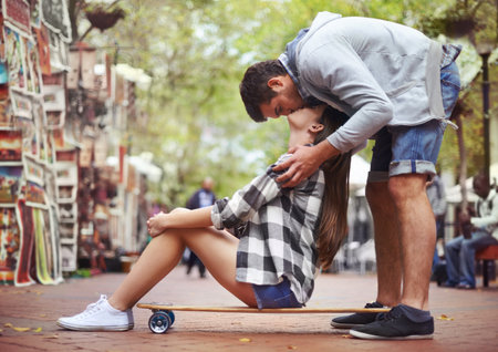 Kisses from above. A young woman sitting on a skateboard outside as her boyfriend stand behind her and kisses her from above.の写真素材