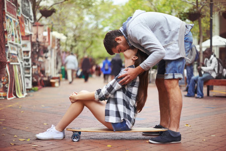 Kisses from above. A young woman sitting on a skateboard outside as her boyfriend stand behind her and kisses her from above.の写真素材