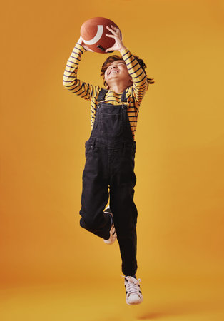 An adorable little boy smiling while catching a football against an orange background. Cute mixed...
