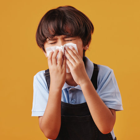 One mixed race Asian boy blowing his nose while looking sick and posing against an orange copyspace background. Cute Asian kid with a runny nose and an allergyの写真素材