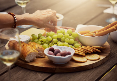Unknown woman taking from a variety of snacks on a tapas wooden board outside. Cheese, bread, fresh grapes and cold meats arranged for lunch on vineyard. Food and wine tasting on farm during a weekenの写真素材