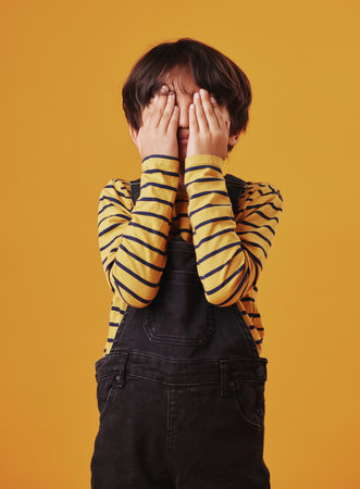 A cute little Chinese boy hiding his face while playing a game and posing against an orange copyspace background. Mixed race child with autism, avoiding and hiding his faceの写真素材