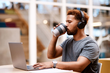 Sipping coffee while making success his. a young businessman wearing headphones and drinking coffee while working on a laptop in an office.の写真素材