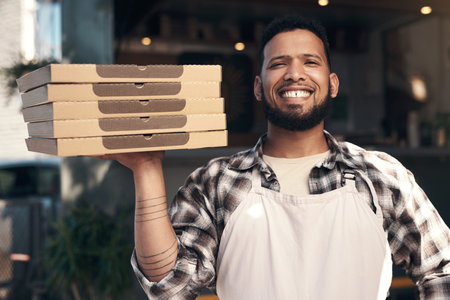 Your pizza is on its way. a handsome young man standing outside his restaurant and holding a stack...