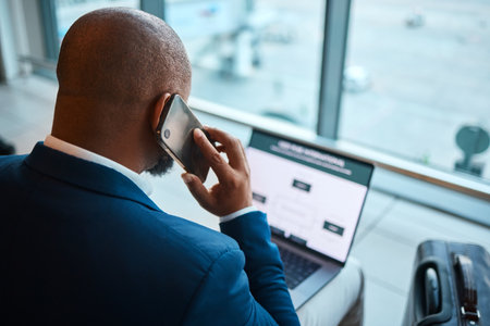 Phone call, laptop and man at airport with travel agency website, schedule and communication services. African business person in waiting room or lobby with suitcase, smartphone and booking onlineの写真素材