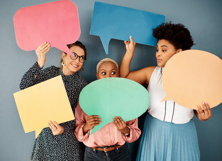 Guys that was supposed to be a secret. Studio shot of a group of attractive young businesswomen holding speech bubbles while standing against a grey background.の写真素材