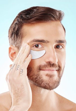 Face, skincare and man with eye patch in studio isolated on a blue background. Beauty, portrait and male model with pad product for dermatology, cosmetics and facial treatment, wellness and health.の写真素材
