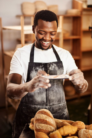 Posting online really helps my business. a male baker taking a picture on his cellphone of a selection of freshly baked bread.の写真素材