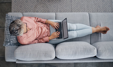 Senior woman, laptop and sofa above relaxed in the living room checking email, typing or writing at home. Elderly female freelancer or writer relaxing on lounge couch working or reading on computerの写真素材