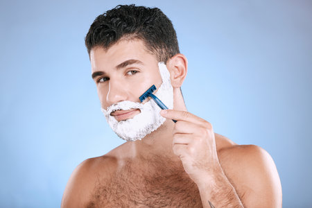 Shaving, grooming and portrait of a man with cream for beard isolated on a blue background. Skincare, beauty and model with razor to shave hair on face with foam for a clean facial look on a backdropの写真素材