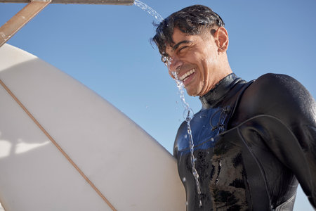 Man, surfing and water shower at beach on face, smile or blue sky in summer, sunshine or ocean travel. Happy surfer cleaning body at tap for sea sports, relax holiday or vacation adventure in wetsuitの写真素材
