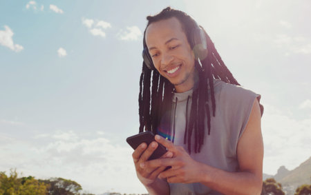 Sky, black man and smartphone with headphones streaming music, online reading and podcast outdoor. African American male, guy and cellphone for social media, headset and listen to audio and soundsの写真素材