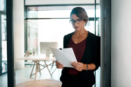 Paperwork is still important in modern business. an attractive young businesswoman standing alone in her office and reading paperwork.の写真素材