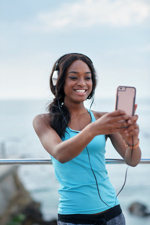 Black woman, phone and headphones at beach, selfie and smile for video call, music or funny meme to...