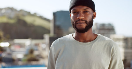 Black man, serious and portrait on rooftop in city, wind or confident in summer on holiday. Young, man and buildings in urban, metro or town with proud, sunshine and beard with focus, vision or dreamの写真素材