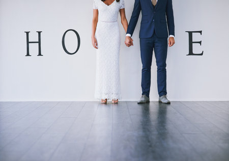 Our story starts here. Concept studio shot of a bride and groom making an M in the word home against a wall.の写真素材