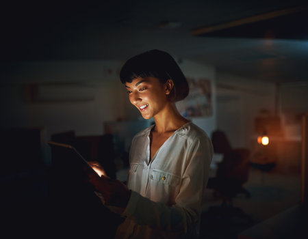 Late night work tasks are a breeze with smart apps. a young businesswoman using a digital tablet during a late night at work.の写真素材