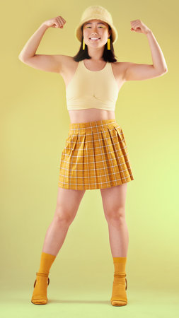 Smile, empowerment and asian woman flexing arms, full body portrait of gen z model with trendy fashion win in studio. Feminism, power and strength, happy and strong girl isolated on yellow backgroundの写真素材