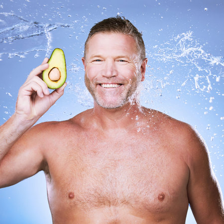 Beauty, water splash with man and avocado for skincare, eco friendly and vegan cosmetics on blue background. Hygiene, hydration and mature male smile in portrait, detox facial and fruit with faceの写真素材