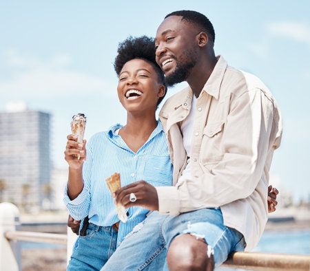 Happy couple at the beach eating an ice cream cone while on a date in nature while on spring vacation. Black man and woman talking and laughing while hugging and having dessert on holiday by seaside.の写真素材
