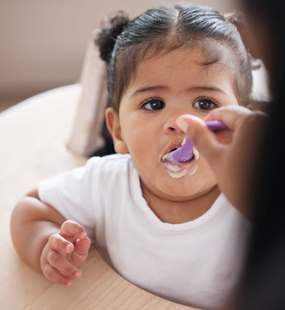 Baby, food and hand feeding with spoon while eating for nutrition, development and growth in a chair. Face of girl toddler or child having breakfast, lunch or dinner while hungry in family homeの写真素材