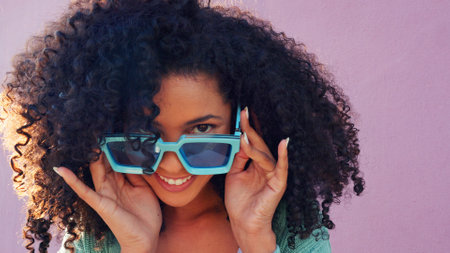 Black woman with sunglasses, smile, laugh and play with hair against pink backdrop in sunshine. Model girl with curly afro hair, happy with blue fashion glasses against pink wall or backgroundの写真素材