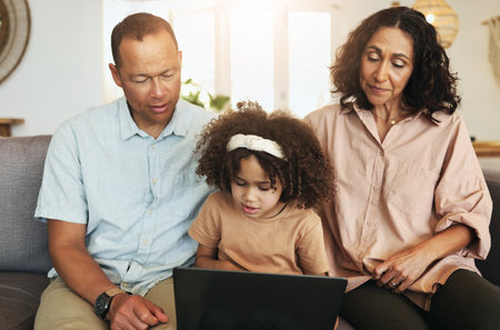 Child streaming a movie on laptop with her grandparents while relaxing on sofa in the living room. Technology, rest and girl watching video or film with grandfather and grandmother for entertainment.の写真素材